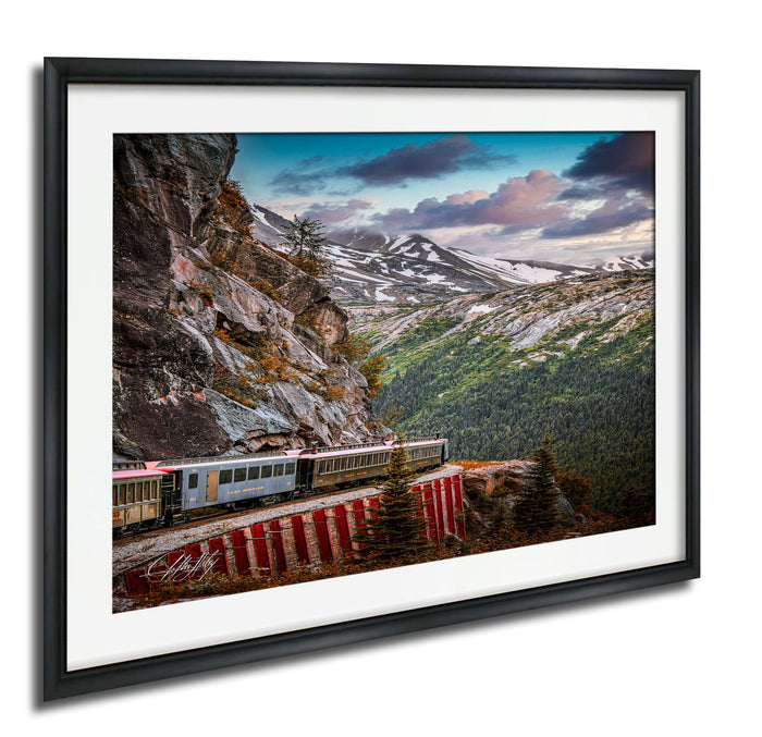 A train travels along a track that runs along the side of a steep, rocky mountain, with snow-capped peaks and a cloudy sky in the background.
