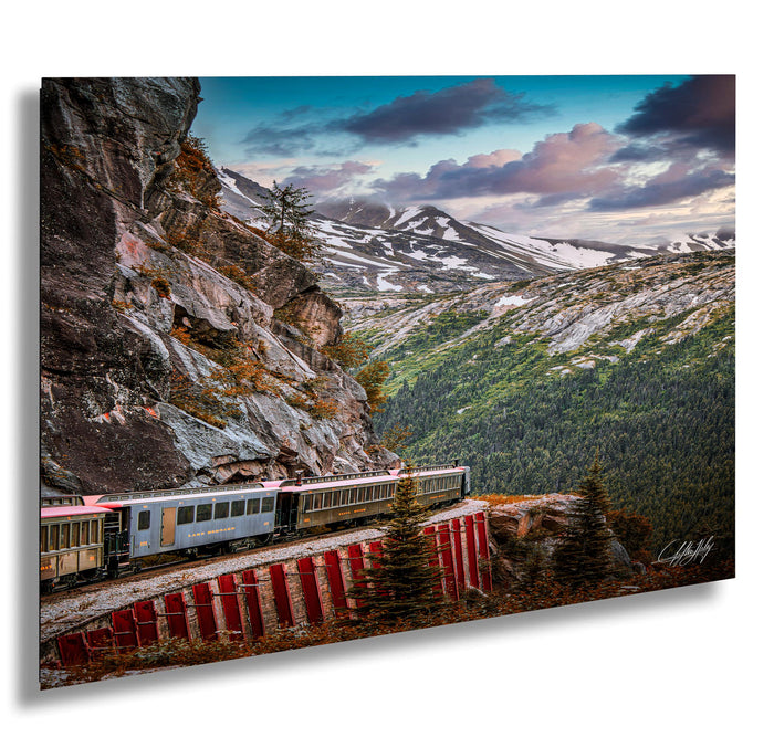 A train travels along a winding track that runs parallel to a steep, rocky cliff face, with snow-capped mountains visible in the background under a partly cloudy sky.
