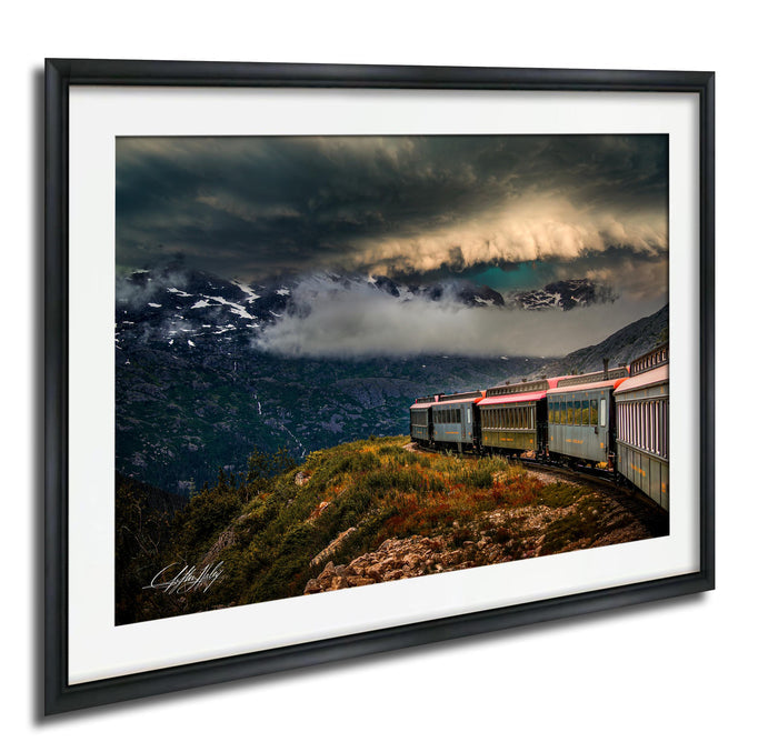 A framed photograph depicting a train traveling through a mountainous landscape with a dramatic cloudy sky in the background.
