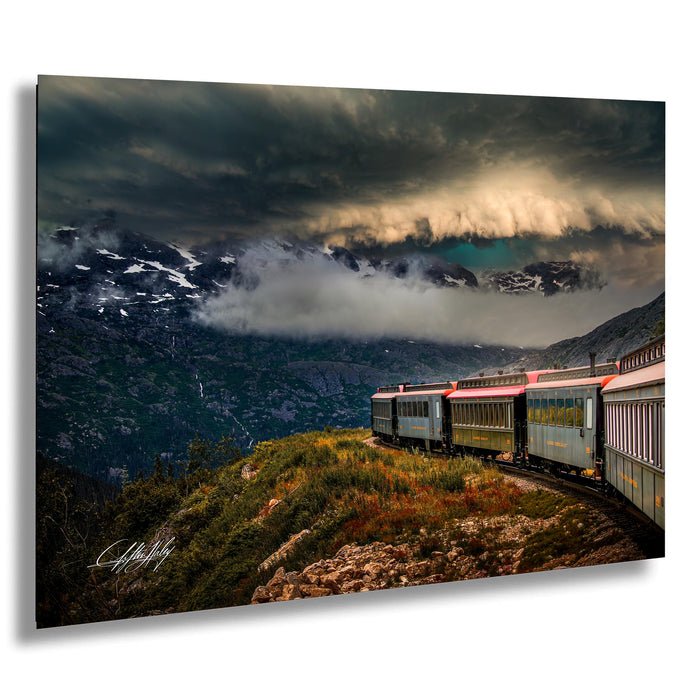 A train travels through a mountainous landscape, with snow-capped peaks and a dramatic cloudy sky in the background.