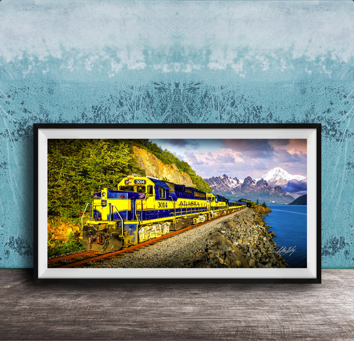 A framed photograph of a yellow and blue train traveling along a track through a mountainous landscape with a body of water in the background.