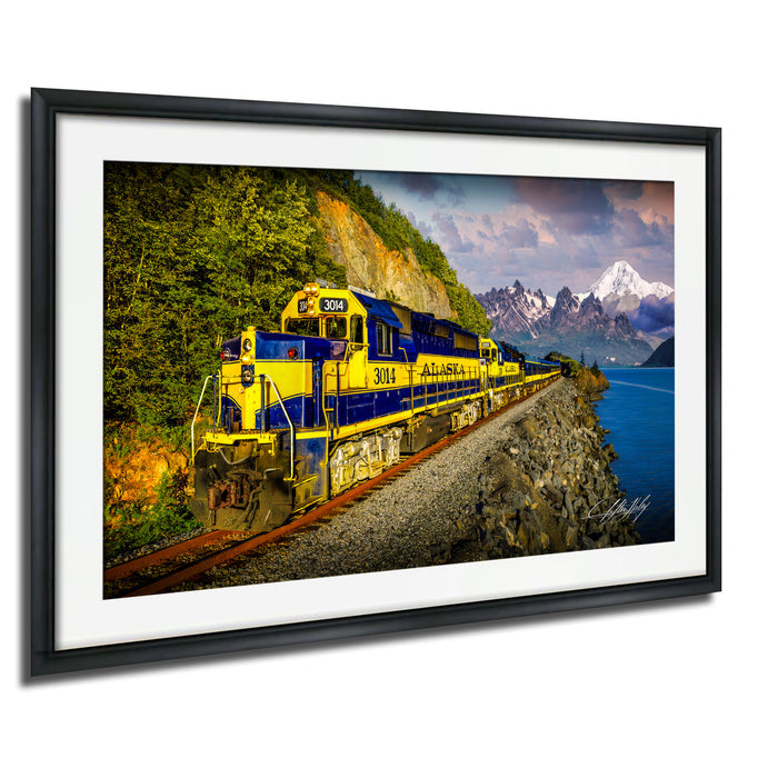 A yellow and blue train engine, numbered 3001, is traveling along a track that runs along the edge of a cliff overlooking a body of water and snow-capped mountains in the distance.