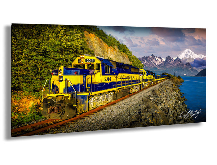 A yellow and blue Alaska train is traveling along a track next to a body of water, with mountains in the background.