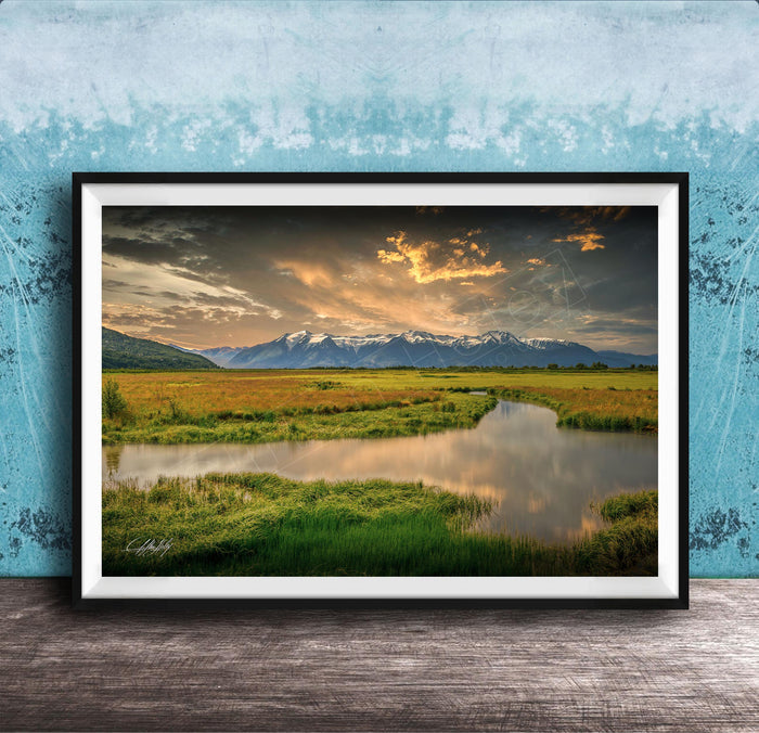 The image is a framed landscape photograph depicting a serene scene of a river flowing through a grassy field, with mountains in the background under a dramatic sky.