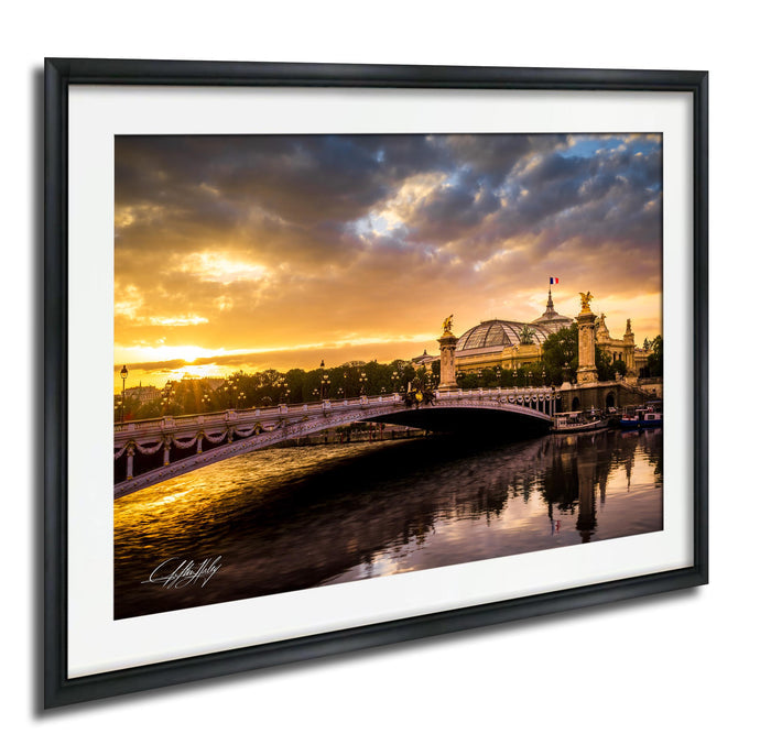 a picturesque scene of a bridge spanning a river, with a large domed building on the other side. The sky is filled with clouds, and the sun is setting, casting a warm glow over the entire scene. The bridge is adorned with statues, adding to its majestic appearance. The image is framed and appears to be a painting or photograph, capturing the beauty of the landmark.