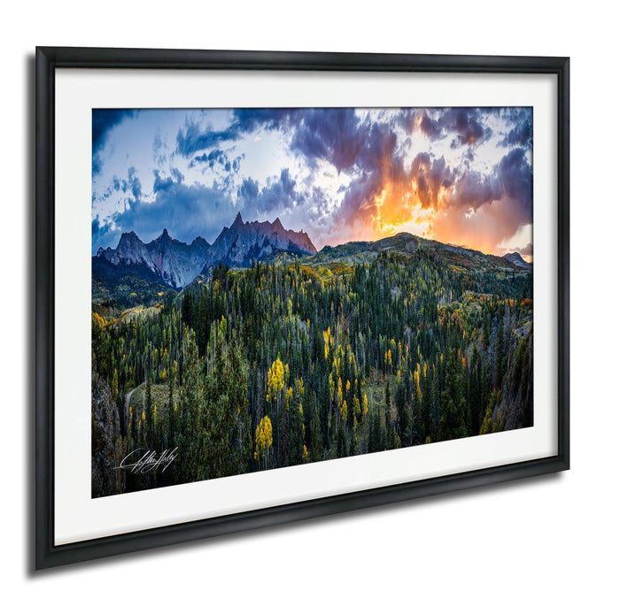 The image is a framed landscape photograph depicting a mountainous, forested area with a vibrant sunset sky in the background.