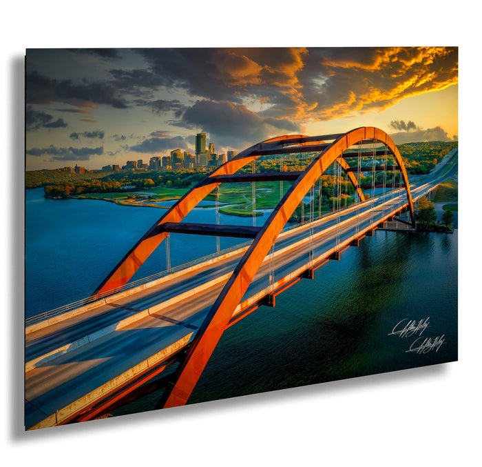 a large, curved bridge spanning across a body of water, with a city skyline visible in the background. The bridge is painted in a striking orange color and has a distinctive arch design. The sky above the scene is filled with vibrant colors, suggesting either sunrise or sunset. The image appears to be a collage of multiple images, with the focus on the bridge and the cityscape.