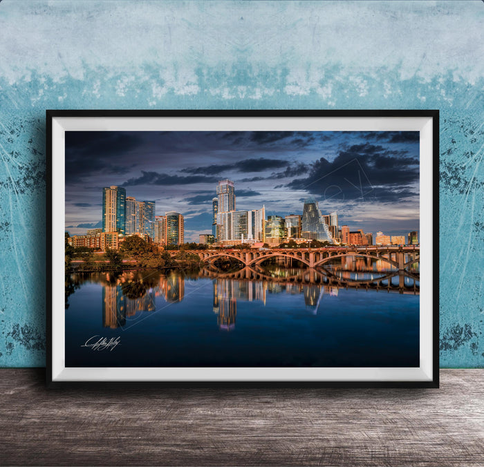 A framed photograph of a city skyline at sunset, with the buildings reflected in the water below.