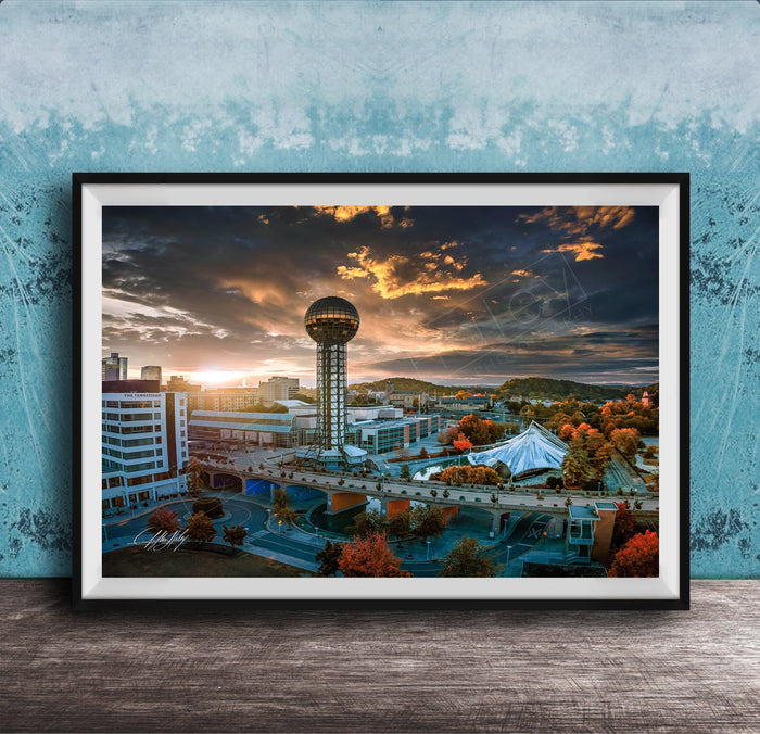The image is a framed photograph of a cityscape at sunset, featuring a tall tower, a bridge, and a large tent.
