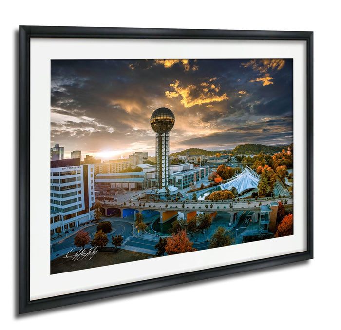 a cityscape with a large tower in the center, surrounded by buildings and a bridge. The sky is filled with clouds, and the sun is setting, creating a beautiful and dramatic atmosphere.