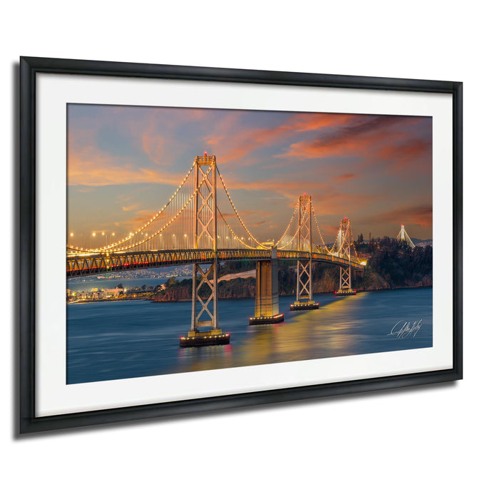 A framed photograph of the San Francisco Bay Bridge at sunset, with the bridge illuminated by warm, golden lights against a vibrant sky.