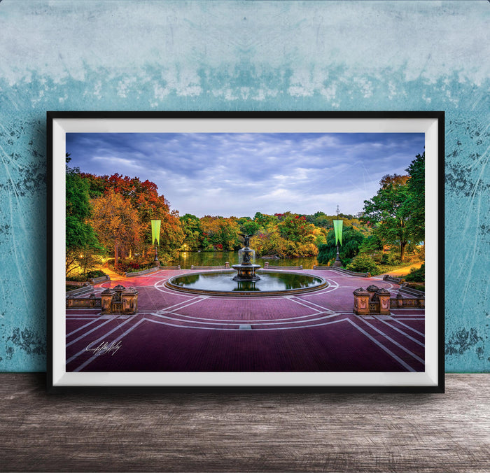 A framed photograph of a park with a fountain, surrounded by trees with autumn foliage, set against a blue wall.