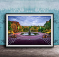 A framed photograph of a park with a fountain, surrounded by trees with autumn foliage, set against a blue wall.