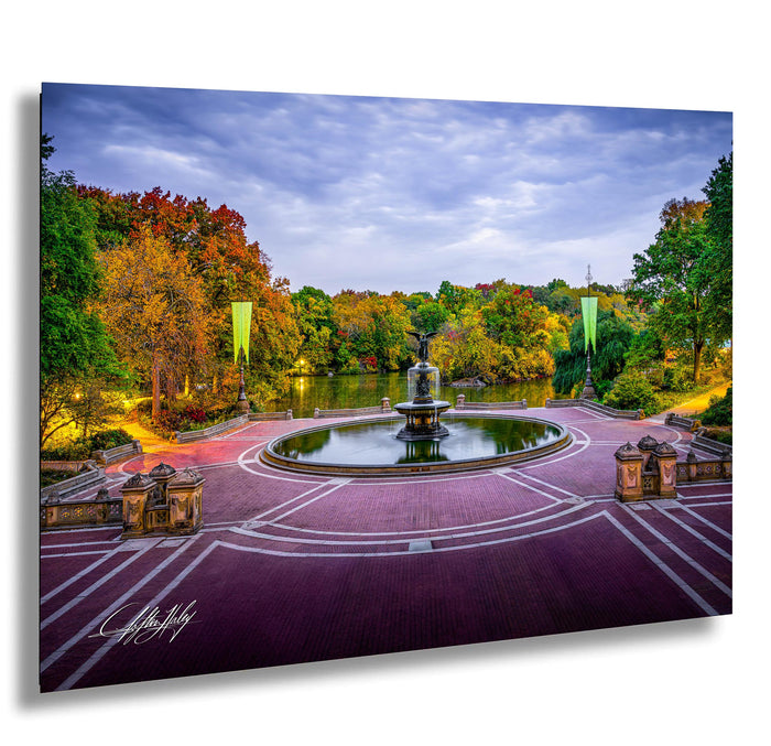 A serene park scene with a circular fountain surrounded by a brick walkway, trees with autumn foliage, and a body of water in the background.