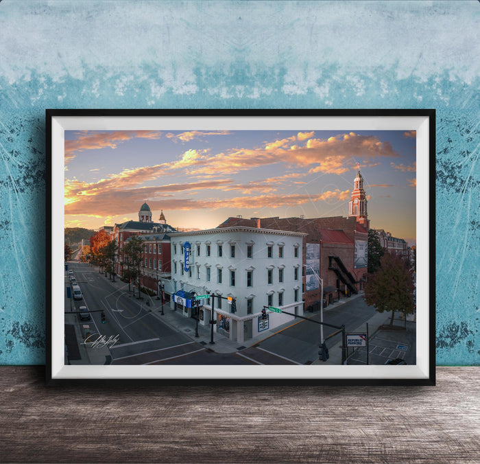 The image is a framed photograph of a city street at sunset, featuring a mix of architectural styles and a church with a tall tower.