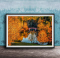 A framed photograph depicting a scenic autumn landscape with a red bridge spanning a tranquil lake surrounded by vibrant orange and yellow foliage.