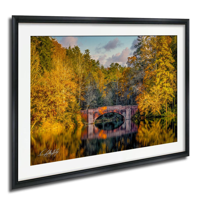 A framed photograph depicting a stone bridge surrounded by a forest with autumn-colored trees reflected in the water below.
