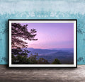 A framed photograph of a scenic mountain landscape at sunset, with a silhouetted tree in the foreground and a hazy, purple sky in the background.