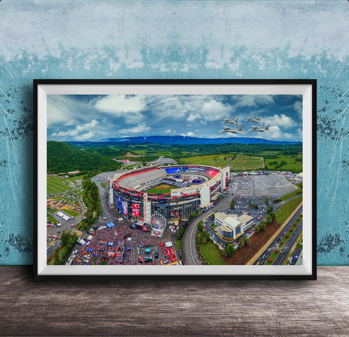 The image is an aerial view of a large stadium surrounded by a green landscape, with a few buildings and vehicles visible.