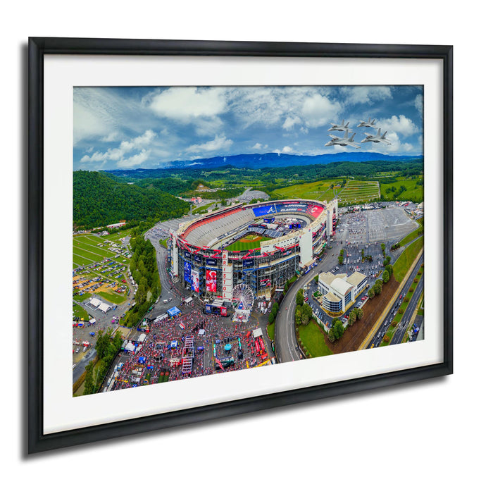 The image is an aerial view of a large stadium surrounded by a large field, with a few buildings and vehicles visible in the vicinity.