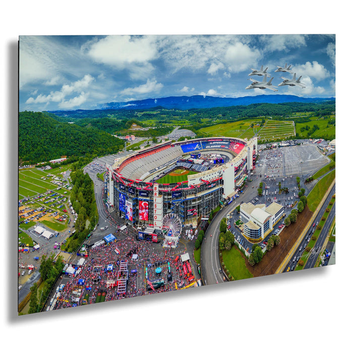 An aerial view of a large sports stadium surrounded by a lush green landscape, with a ferris wheel and other attractions visible. Several planes can be seen flying in the sky above the stadium.