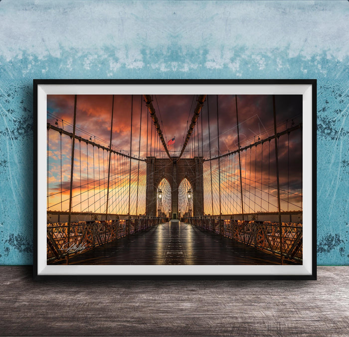 A framed photograph of the Brooklyn Bridge at sunset, with the bridge's intricate metal structure and suspension cables silhouetted against a vibrant sky filled with clouds and warm hues.