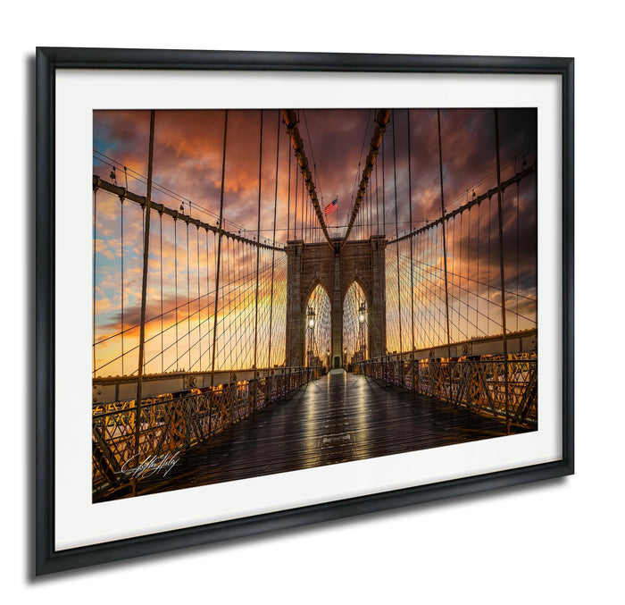 A framed photograph of the Brooklyn Bridge at sunset, with the iconic stone towers and suspension cables illuminated against a vibrant sky.