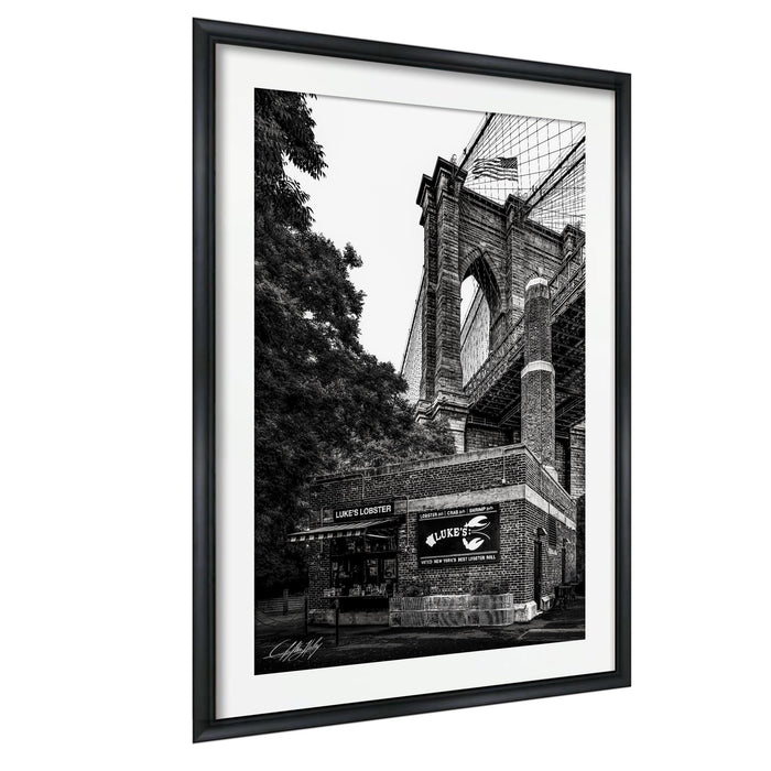 A black and white photograph of the Brooklyn Bridge, with the American flag flying atop the structure.