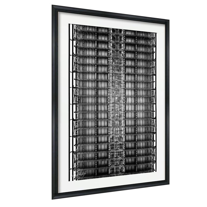 The image is a black and white photograph of a tall, intricate metal structure with numerous levels and rows of bars or beams. The photograph is framed and displayed on a wall.
