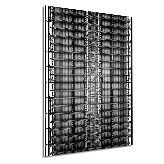 The image is a black and white photograph of a tall, intricate metal structure with a grid-like pattern of beams and supports.