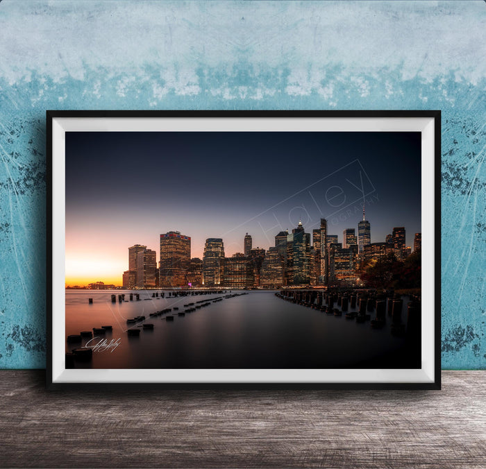 A framed photograph of the skyline of New York City at sunset, with the city's skyscrapers and landmarks illuminated against a dark blue sky.