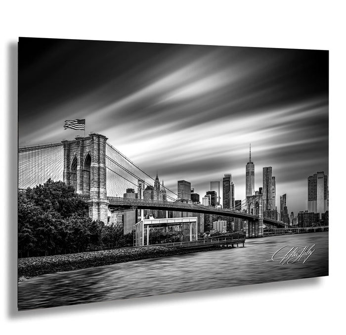 A black and white photograph of the Brooklyn Bridge and the New York City skyline, with the bridge spanning across the frame and the city's skyscrapers visible in the background.