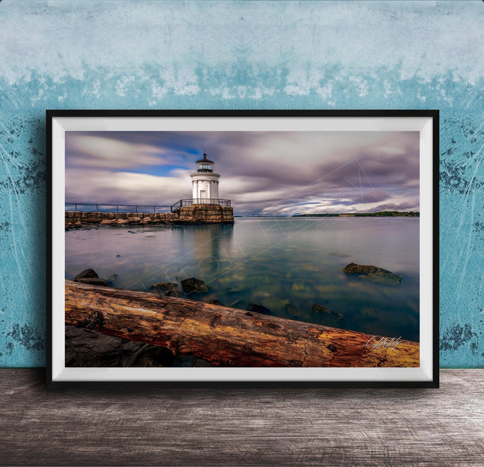 A framed photograph of a lighthouse on a rocky shore, with a calm body of water in the background and a cloudy sky above.
