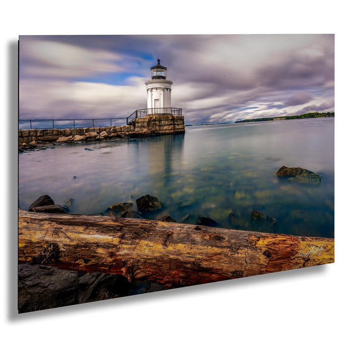 A white lighthouse stands on a rocky shore, overlooking a calm body of water with a cloudy sky in the background.