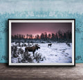 A framed photograph depicting two moose standing in a snowy field with a forest in the background, under a pink and purple sky.