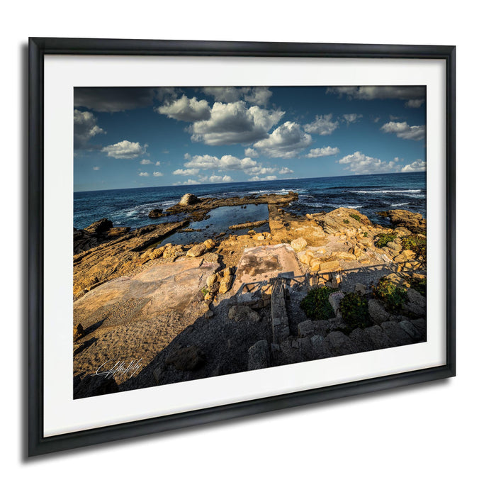 The image is a framed photograph of a rocky coastal landscape with a clear blue sky and white clouds.