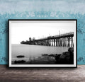 A black and white photograph of a long pier extending into the water, with a rocky shoreline in the foreground.