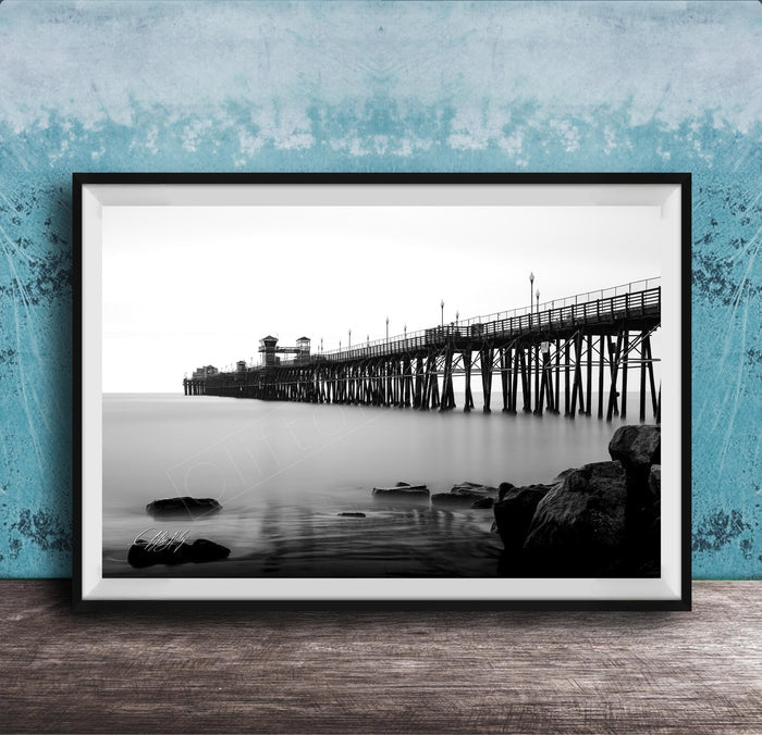 A black and white photograph of a long pier extending into the water, with a rocky shoreline in the foreground.