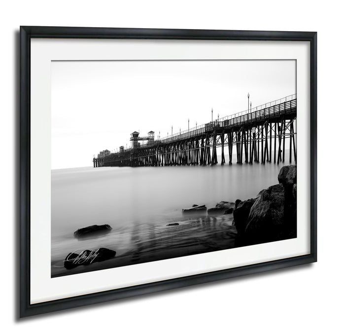 A black and white photograph of a long pier extending into the water, with rocks and a calm body of water in the foreground.