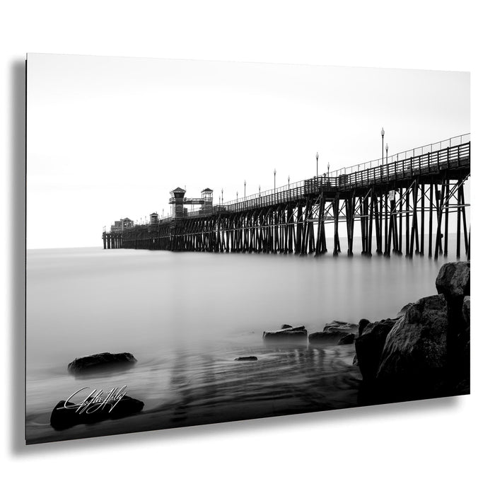 A black and white photograph of a long pier extending into the water, with a rocky shoreline in the foreground.