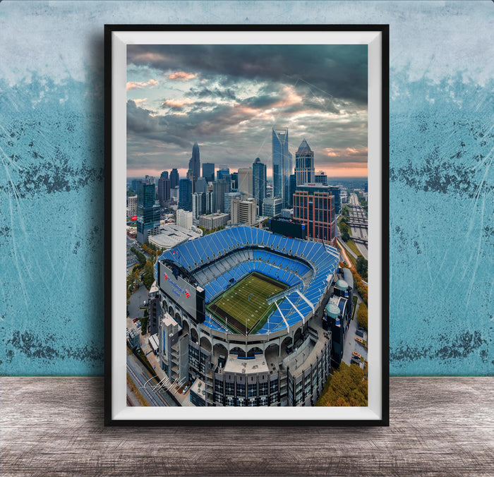 An aerial view of a large sports stadium with a city skyline in the background, framed in a black frame.