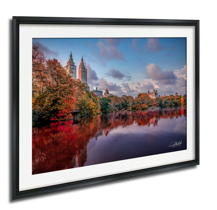 a serene cityscape with a calm lake reflecting the surrounding buildings and trees. The buildings have a mix of architectural styles, and the trees are adorned with autumn foliage, creating a picturesque scene.