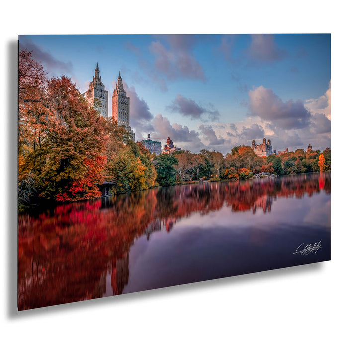 a serene cityscape with a calm lake reflecting the vibrant colors of autumn foliage. The tall buildings in the background are bathed in warm hues, creating a picturesque scene. The sky above is a clear blue, dotted with fluffy white clouds, adding to the overall beauty of the image. The composition of the photograph, with the lake and the buildings in the foreground and the sky in the background, creates a sense of depth and perspective.