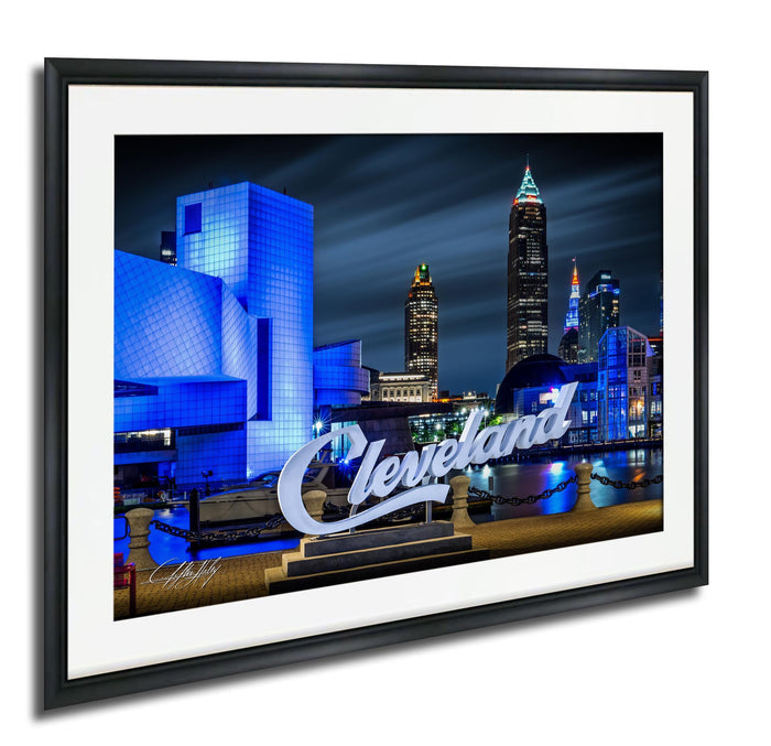 A framed photograph of the Cleveland skyline at night, with the city's iconic buildings illuminated in blue and white lights.