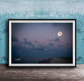 A framed photograph of a serene beach at night, with a full moon rising over the ocean and a cloudy sky.