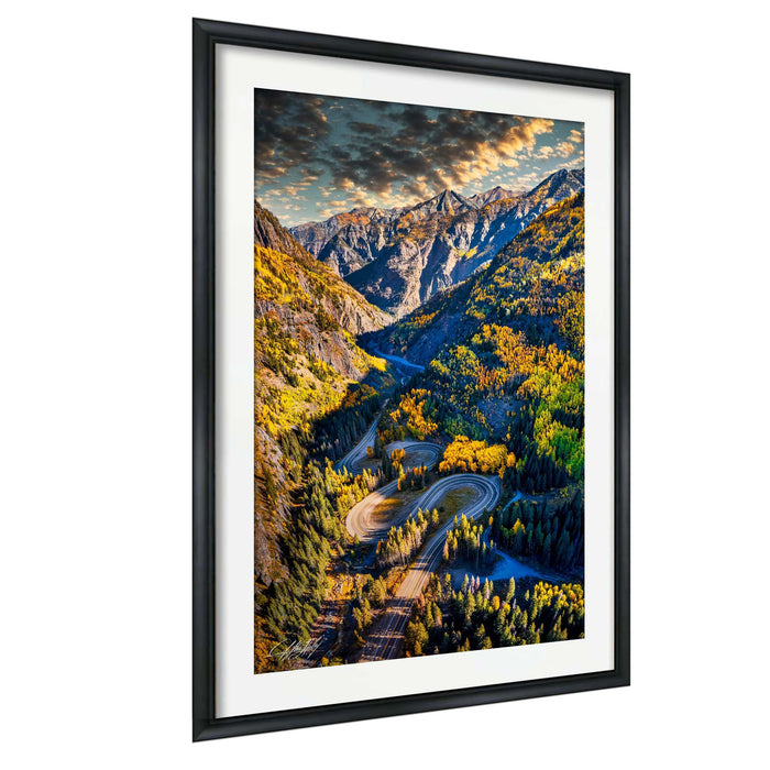The image is a framed photograph depicting a winding mountain road surrounded by lush green forests and autumnal trees.