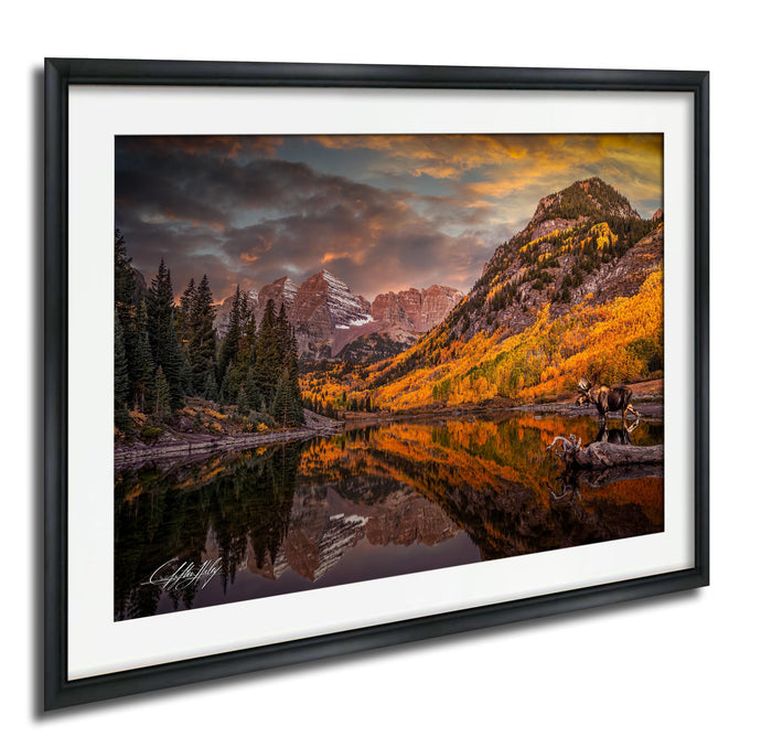 a serene mountain landscape with a lake reflecting the vibrant colors of the autumn foliage. The mountains are adorned with a mix of green and orange hues, creating a picturesque scene. The sky above is filled with clouds, adding to the dramatic atmosphere of the landscape. In the foreground, there are two moose standing near the lake, adding a touch of wildlife to the scene. The image is framed and appears to be a painting or photograph, capturing the beauty of nature.