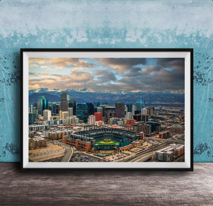 The image is a framed aerial view of a city skyline, featuring a baseball stadium and mountains in the background.
