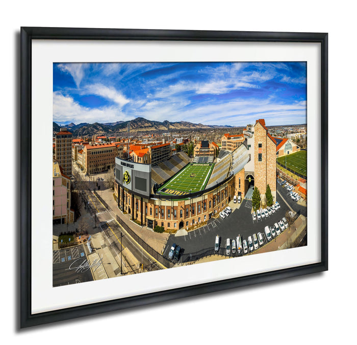 The image is a framed aerial view of a large sports stadium, likely a football stadium, surrounded by a cityscape with buildings and a parking lot.