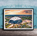 The image is a framed painting depicting a large stadium with a curved roof, set against a backdrop of a city skyline and a colorful sunset sky.